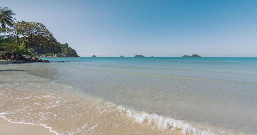 Skaista pludmales aina ar mierīgu ūdeni un attālinātiem salām, kas redzamas horizontā. Zaļi koki malā papildina idillisko skatu.