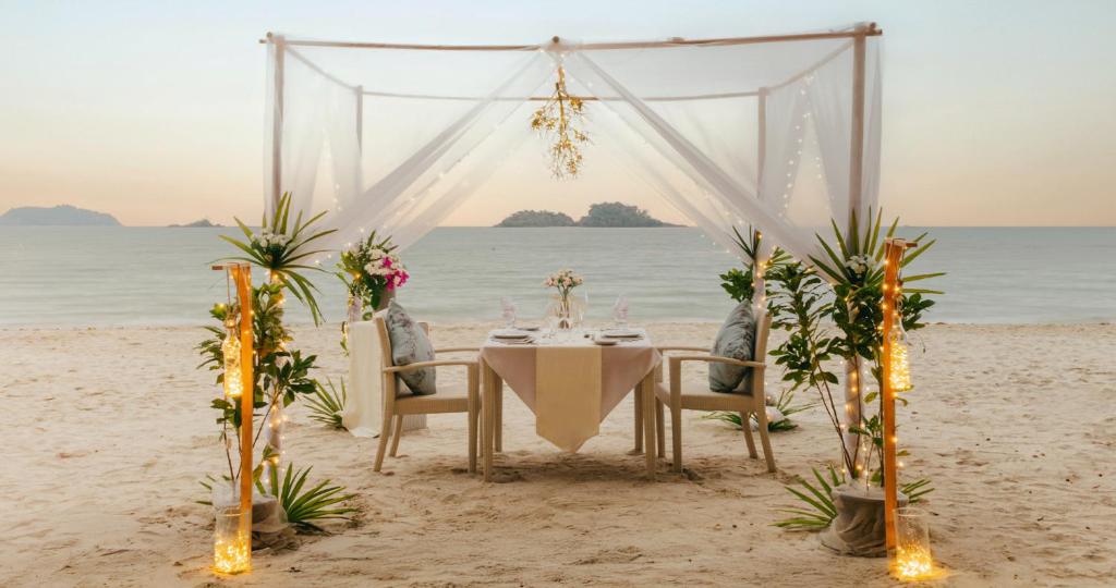 Romantisks galda iestatījums pludmalē, apņemts ar augiem un gaismiņām, kas rada burvīgu atmosfēru saulrieta laikā.