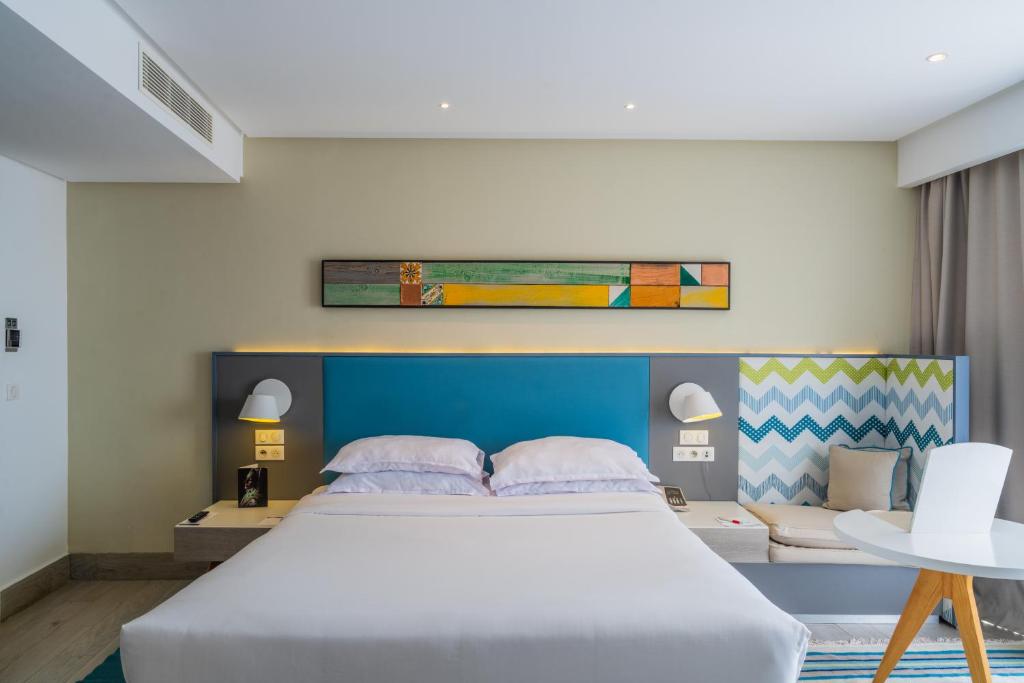 Mūsdienīga guļamistaba ar spilgtu gultas galvu un krāsainu mākslas darbu pie sienas, kas piešķir telpai stilīgu un komfortablu atmosfēru. Gultas blakus ir ērta atpūtas zona un galdiņš.