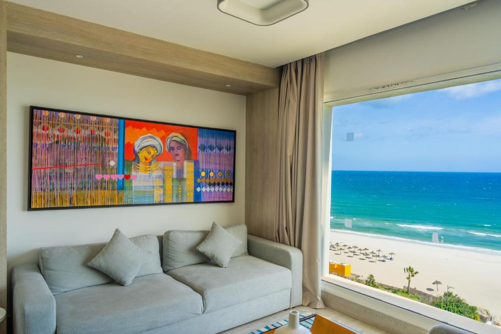 Mūsdienīga viesistaba ar lielu logu, caur kuru redzams pludmales skats, un krāsaina glezna uz sienas, kas attēlo divas sievietes. Eleganta dīvāna ar spilveniem klātbūtne rada mājīgu atmosfēru.