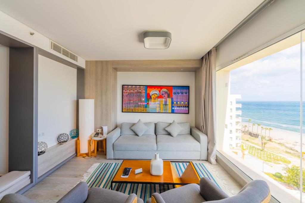 Mūsdienīgs viesistabas interjers ar plašu skatu uz jūru, kurā redzams dīvāns un krēsli, kā arī krāsains mākslas darbs uz sienas.
