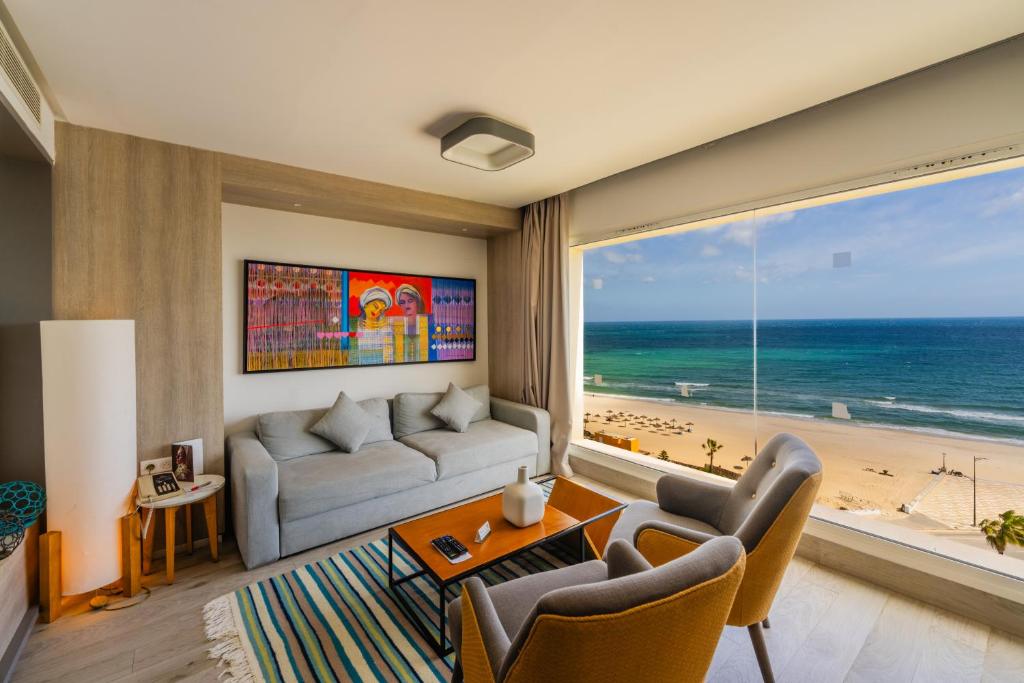 Mūsdienīgs viesnīcas numurs ar lielu logu, kas sniedz skatu uz pludmali un jūru. Interjerā ir stilīgs dīvāns, koka galds un krēsli, kā arī krāsains mākslas darbs pie sienas.