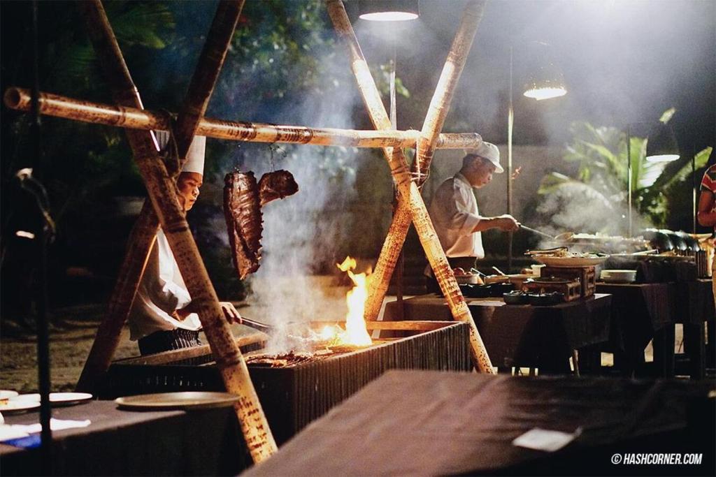 Grilēšanas pasākums naktī, kur divi pavāri gatavo ēdienus uz uguns, radot patīkamu atmosfēru. Dūmi un gaisma piešķir noskaņu, izceļot gardos ēdienus.