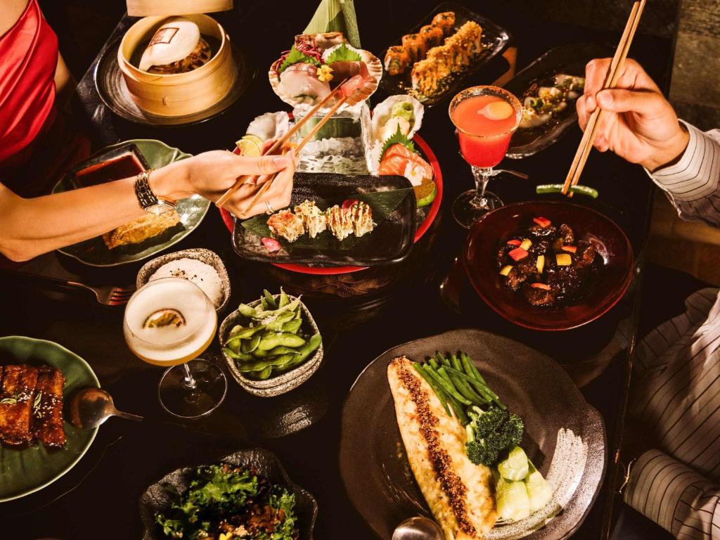 Bagātīgs ēdienu galda klājs ar dažādiem Austrumu ēdieniem, tostarp sushi, dārzeņiem un kokteiļiem, kas tiek baudīti kopā ar draugiem.