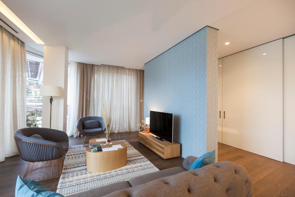 Mūsdienīgs viesistabas interjers ar ērtām mēbelēm, televizoru un gaišām aizkariem, radot mājīgu atmosfēru. Dabiskais apgaismojums piešķir telpai svaigumu.