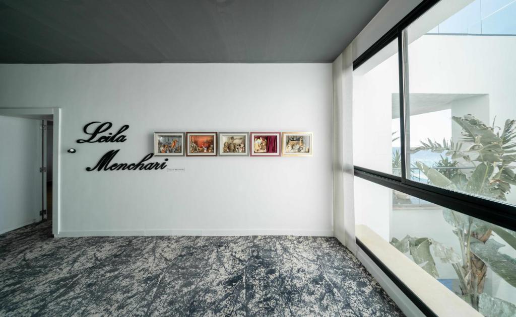 Mūsdienīga izstāžu telpa ar Leilas Menčari nosaukumu un četriem rāmjiem, kuros redzamas gleznas, kas izkārtotas uz baltas sienas blakus liela loga.