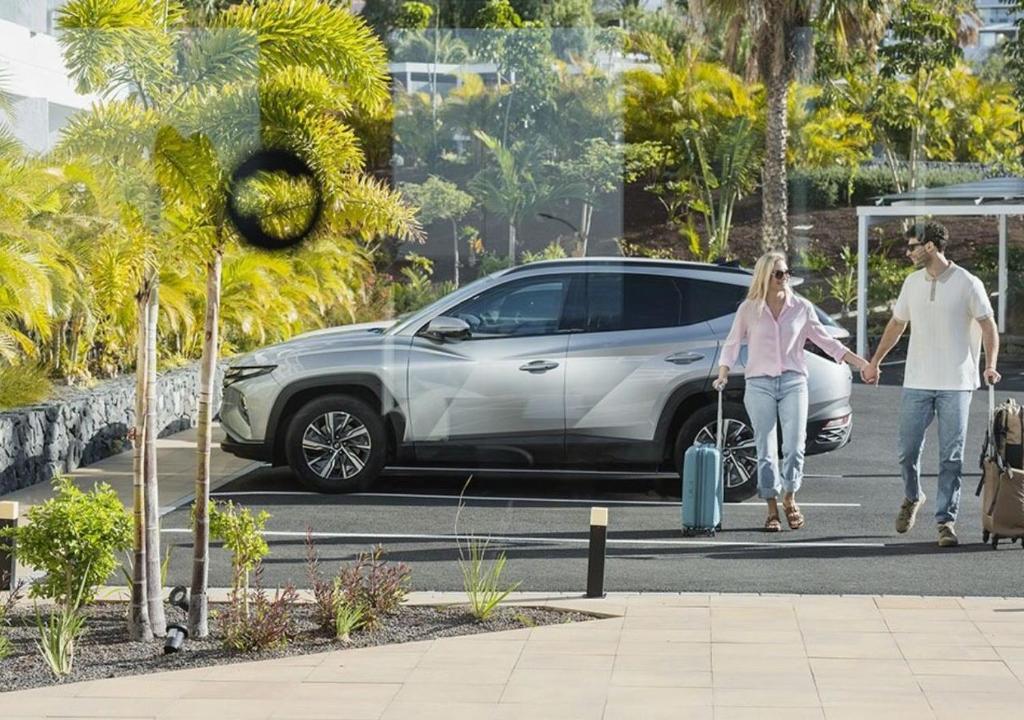 Pāris, turēdami rokās bagāžu, staigā uz auto stāvvietu, kur atrodas sudraba krāsas SUV. Apkārt redzami tropiski augi un zaļa vide, kas rada relaksētu atmosfēru.