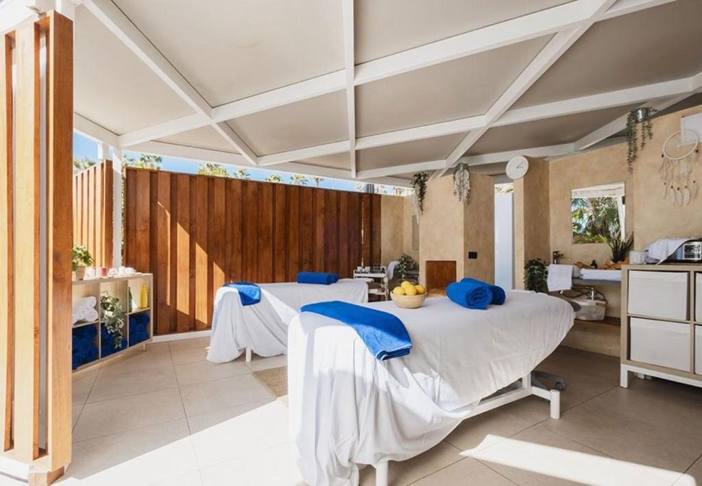 Relaksējoša spa telpa ar divām masāžas galdiem, baltām segām un zilu dvieli. Interjers ir gaišs ar koka akcentiem un augiem, radot mierīgu atmosfēru.