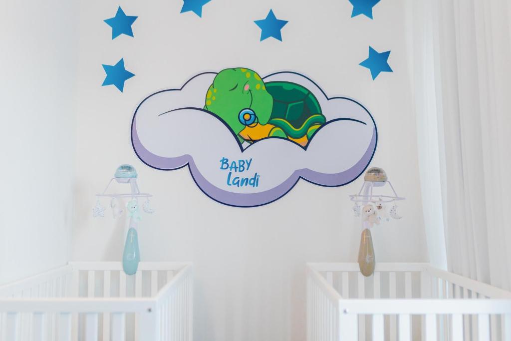 Bērnu istabas interjers ar rotaļu gultiņām un krāsainu mākoņiem uz sienas, uz kuriem attēlots zaļš bruņurupucis un uzraksts "BABY Land". Zvaigznes pie sienas piešķir telpai mīkstu un sapņainu atmosfēru.