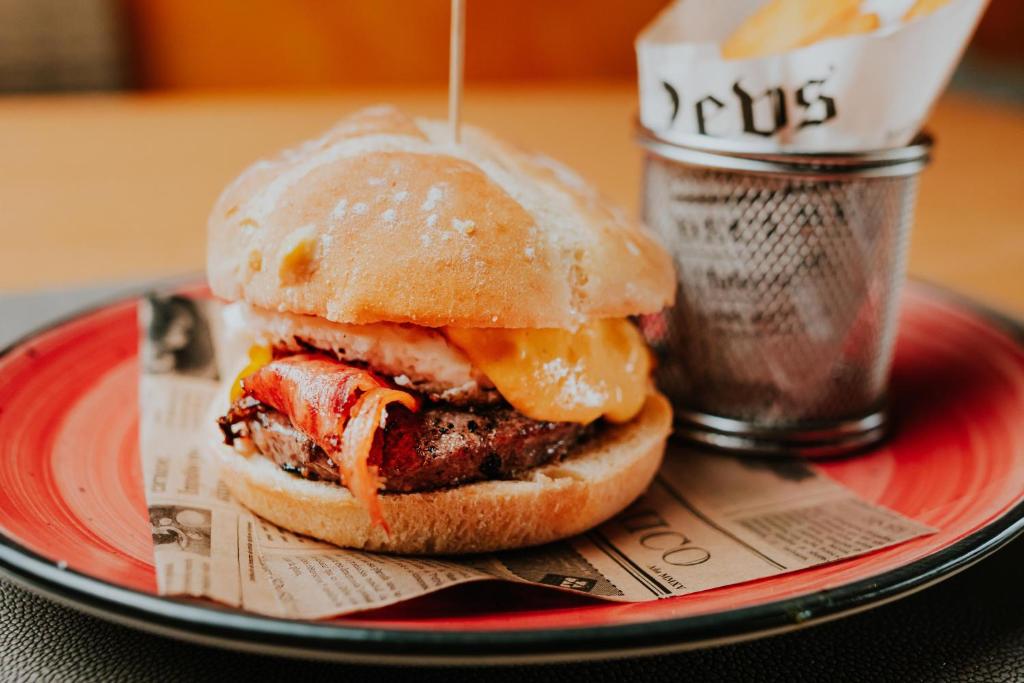 Gards burgeris ar gaļas kotleti, olu un sieru, pasniegts uz sarkanas šķīvja kopā ar frī kartupeļiem.