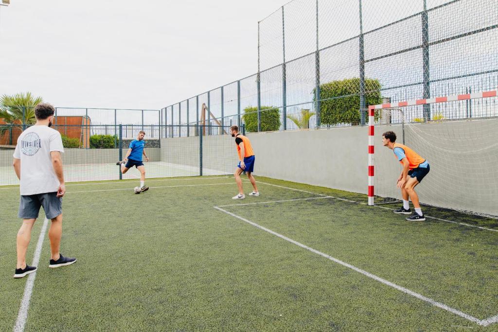Jaunieši spēlē futbolu uz mākslīgā zālāja laukuma, aktīvi piedaloties spēlē un izpildot sitienus. Apkārt ir futbola vārti un svars, kas piešķir dinamiku sporta aktivitātēm.