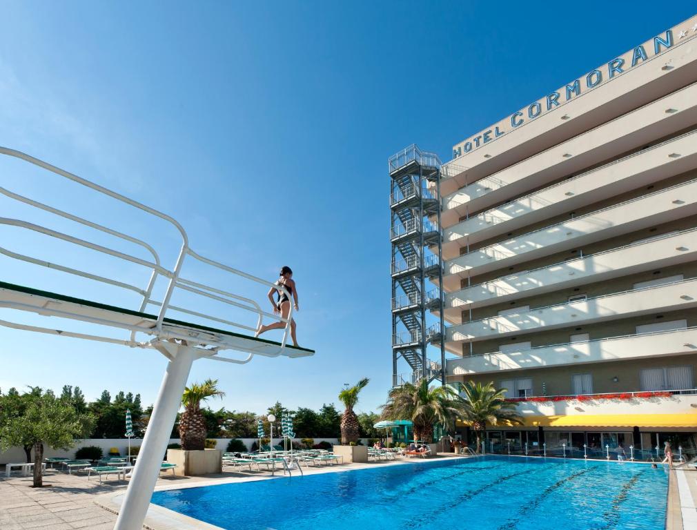 Baseins ar lēcēju un viesnīcu "Cormoran" fons, brīnišķīgs vasaras diena. Viesi atpūšas ap baseinu, kamēr kāds gatavojas lēkt.
