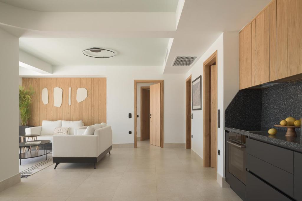 Mūsdienīga viesistaba ar gaišām krāsām, elegantu dīvānu un koka akcentiem, kas rada siltu atmosfēru. Virtuve ir aprīkota ar tumšām mēbelēm un augļiem uz galda.