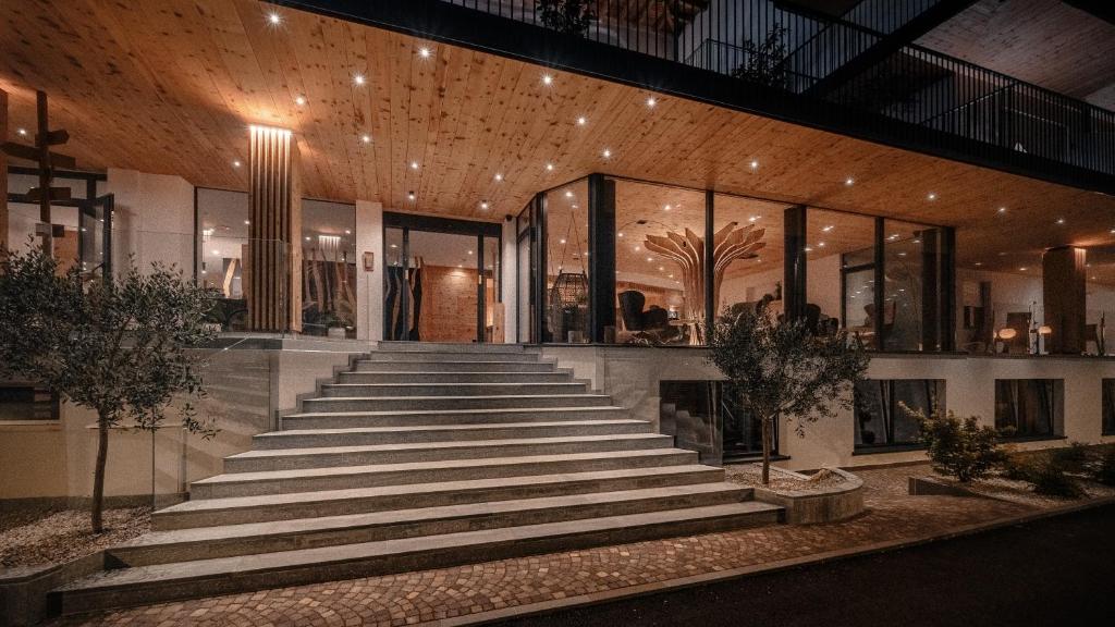 Mūsdienīga arhitektūra ar plašu ieeju un kāpnēm, kas ved uz elegantu interjeru, apgaismotu ar siltu gaismu. Apkārt esošās augu kompozīcijas piešķir dabīgumu un harmoniju.