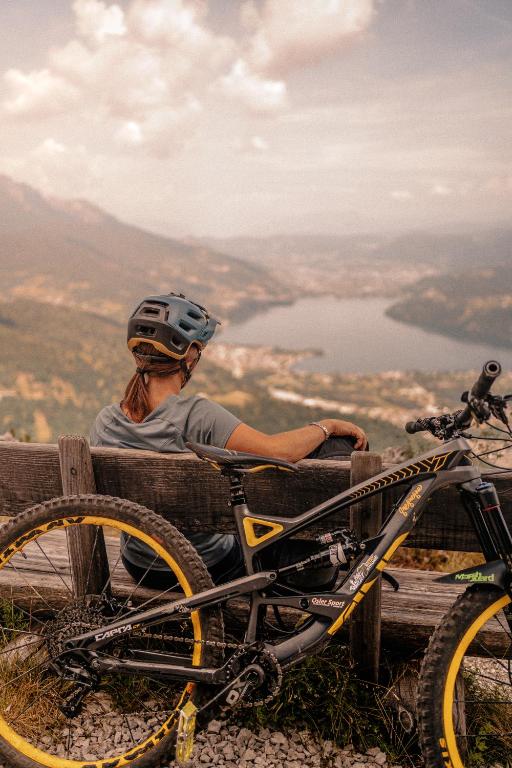 Kļūdas un relaksācijas brīdis kalnu ainavā, kur sportists ar velosipēdu izbauda skatu uz ezeru un apkārtējiem pakalniem.