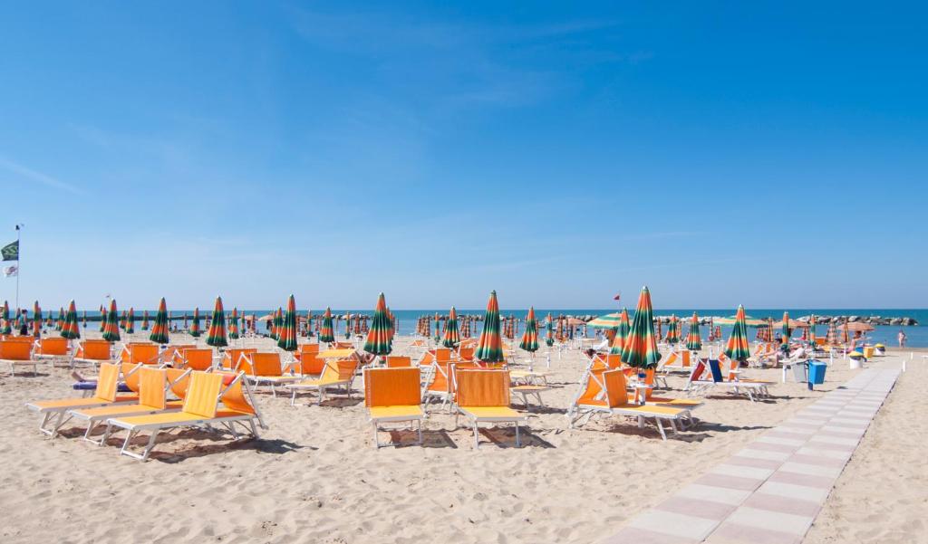 Pludmale ar oranžiem sauļošanās krēsliem un zaļi-oranžām saulessargiem, zem skaidras debess un blakus mierīgai jūrai. Ideāla vieta atpūtai un saules baudīšanai.