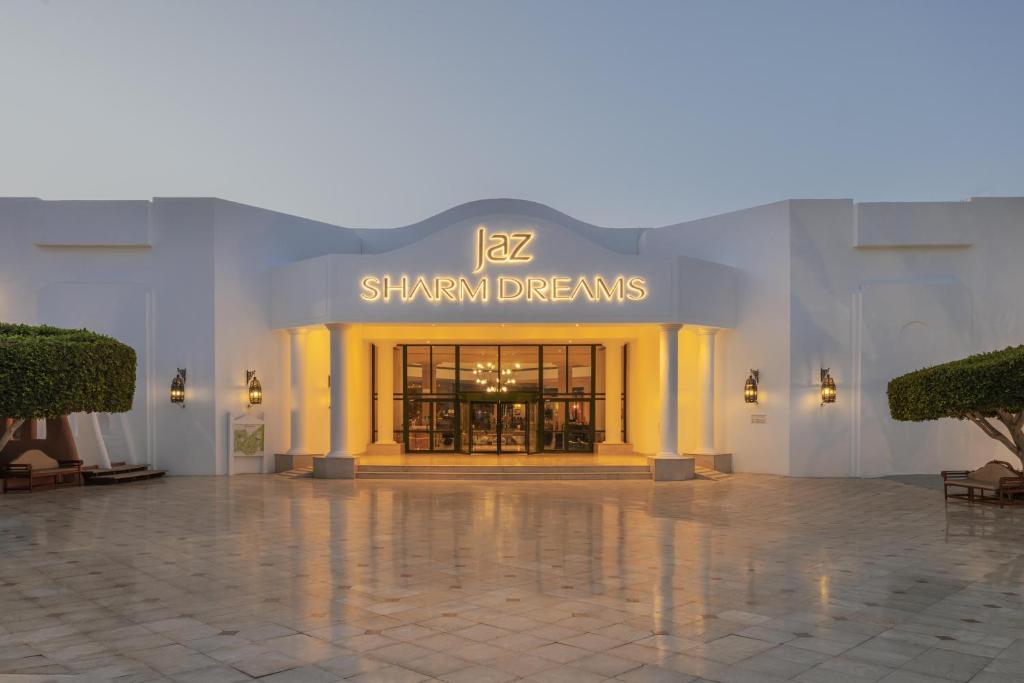 Jaz Sharm Dreams viesnīcas ieeja ar izcili apgaismotu nosaukumu un elegantu arhitektūru. Apkārtne ir sakopta ar zaļiem krūmiem un akmens grīdu, radot viesmīlīgu atmosfēru.