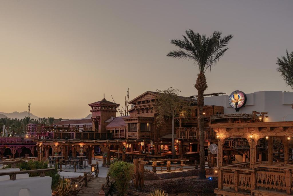 Pilsētas ainava ar tradicionālu arhitektūru un palmu kokiem, kas peld saulrieta gaismā. Izceļas koka struktūras un restorānu terases, radot viesmīlīgu atmosfēru.