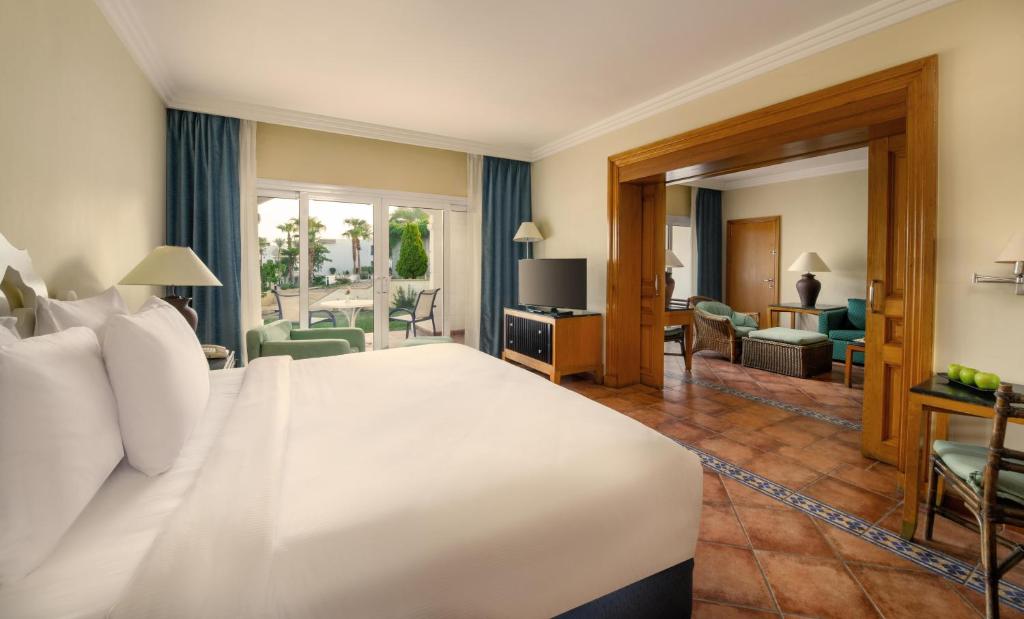 Liela un gaiša viesnīcas istaba ar ērtu gultu, televizoru un piekļuvi terasei, kurā redzami augi un mēbelēti sēdvietas.