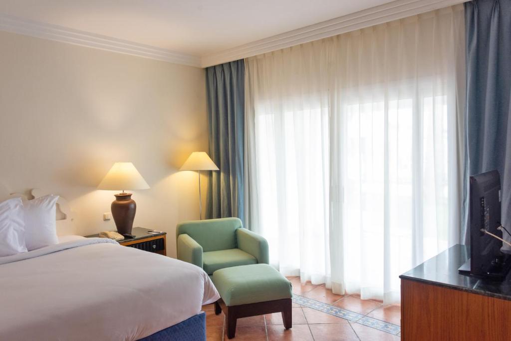 Mūsdienīgs viesnīcas numurs ar ērtu gultu, krēslu un televizoru, kas izgaismots ar dabīgu gaismu no logiem.