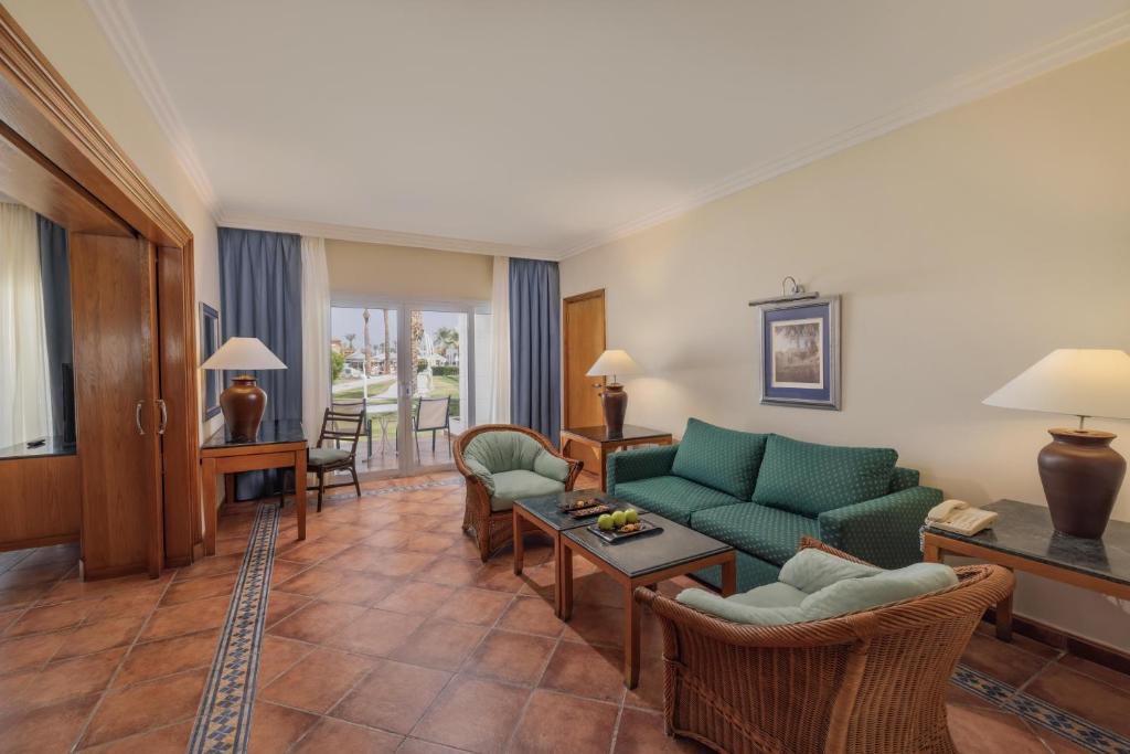 Mūsdienīga viesnīcas viesistaba ar zaļu dīvānu, ērtām krēslām un galdu, uz kura atrodas augļi. Logi vērsti uz āru, ļaujot ieplūst dabiskajai gaismai.