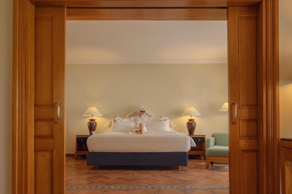 Luksusīgs viesnīcas numurs ar plašu gultu un relaksējošu atmosfēru, redzams caur atvērtām durvīm. Interjerā ir siltas krāsas un eleganti apgaismojumi.