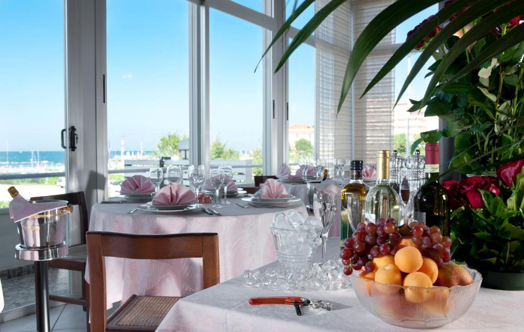 Eleganta restorāna galda iestatījums ar skatu uz jūru, dekorēts ar rozā galdautu, glāzēm un augļu trauku. Apkārt ir ziedi un dzērieni, radot svinīgu atmosfēru.