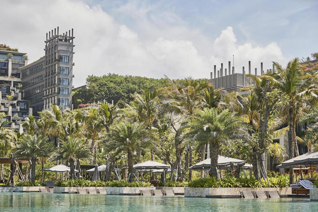 Mūsdienīgs kūrorts ar palmām un peldbaseinu, kas atrodas blakus modernai ēkai, apkārt skaista ainava.