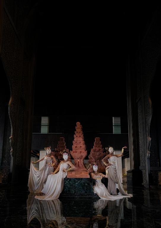 Četras dejotājas tradicionālās drēbēs ar maskām, pozē pie statujas tumšā telpā, radot dramatisku atmosfēru.