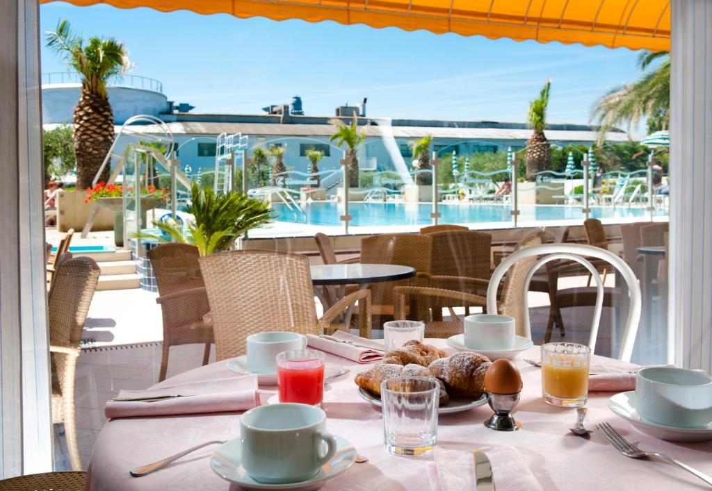 Broka ar skatu uz baseinu, kurā redzami palmām un viesnīcas terasei. Galda klājumā ir kafija, sulas un kruasāni, radot viesmīlīgu atmosfēru.