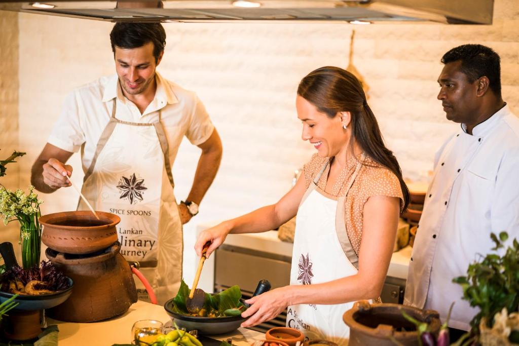 Divi cilvēki gatavo ēdienu, izmantojot tradicionālos katlus, kamēr trešais cilvēks novēro. Visi ir apģērbušies priekšautos, un apkārt ir dažādi dārzeņi un garšvielas.