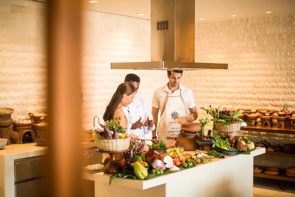 Kulinārijas nodarbība ar šefpavāru, kas sniedz padomus un demonstrē gatavošanu, apkārt bagātīgiem dārzeņiem un augļiem. Vides elements veido siltu un viesmīlīgu atmosfēru.