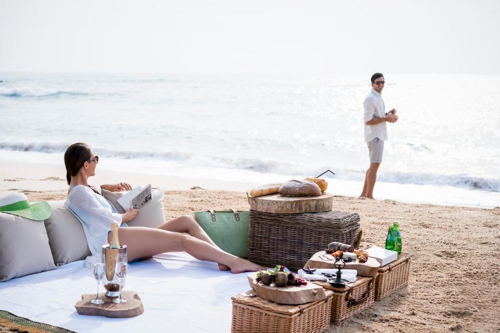 Pludmales ainava ar sievieti, kas lasa grāmatu, un vīrieti, kas stāv pie ūdens. Apkārt ir piknika grozi ar uzkodām un dzērieniem.