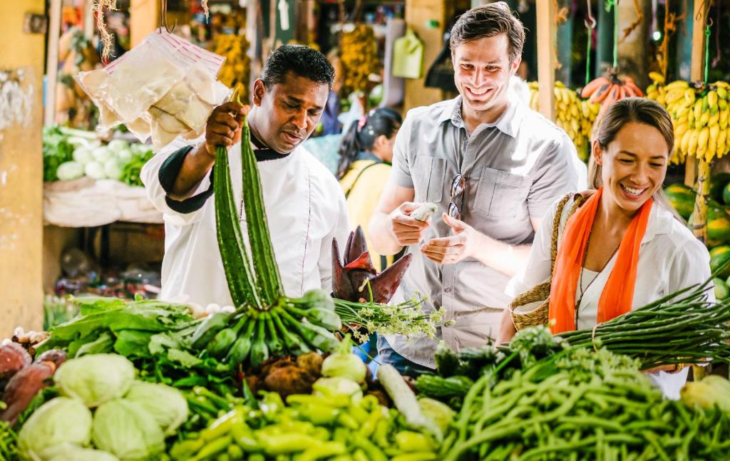 Tirgū notiek dzīvīga saruna, kurā viens no tirgotājiem piedāvā svaigus dārzeņus diviem apmeklētājiem, kuri izskatās priecīgi un ieinteresēti. Fons ir pilns ar krāsainiem augļiem un dārzeņiem.