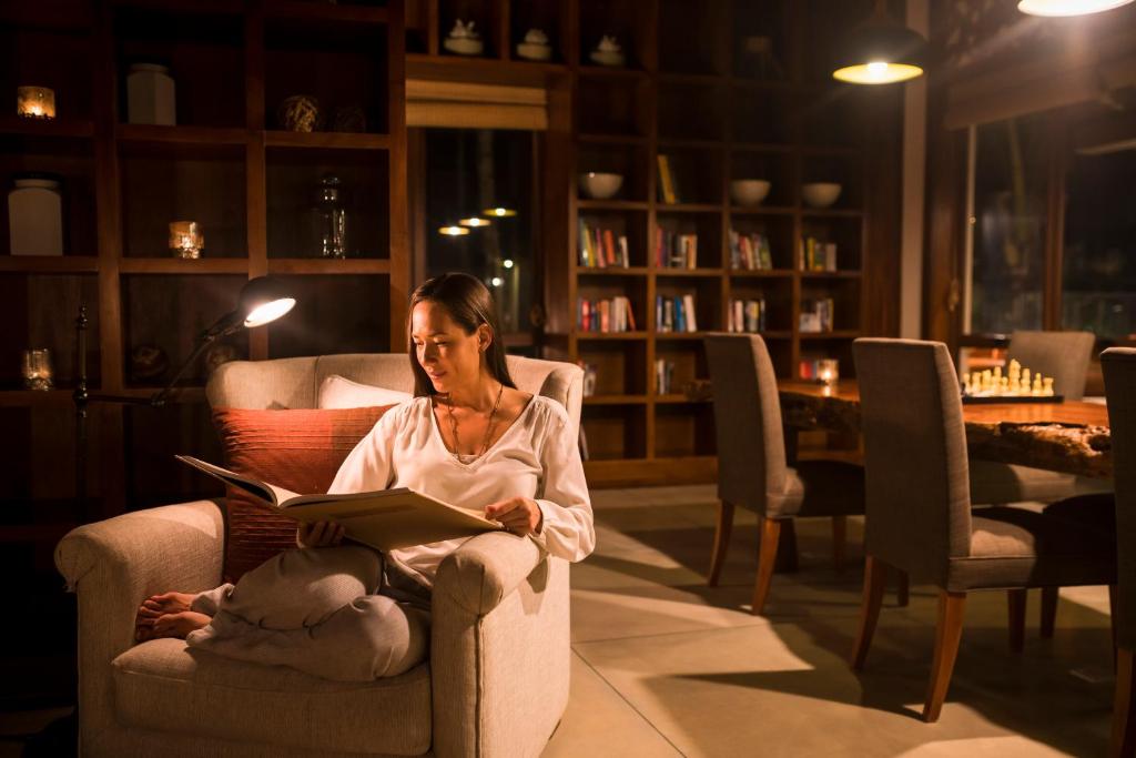 Sieviete, kas ērti sēž krēslā un lasa grāmatu, apgaismota ar galda lampu, ar grāmatu plauktiem un galdu fonā, kur ir šaha galds.