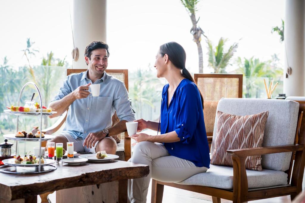 Pāris bauda kafiju un sarunājas eleganti iekārtotā telpā ar dabas skatu. Uz galda ir dažādi gardumi un dzērieni, radot patīkamu atmosfēru.