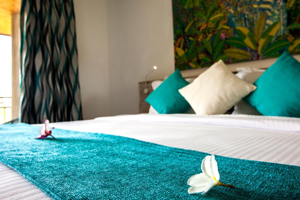 Mūsdienīga guļamistaba ar zaļu gultas pārklāju, spilveniem un ziediem, kas piešķir mājīgu atmosfēru. Aiz loga redzama krāsaina māksla un stilīgas aizkari.