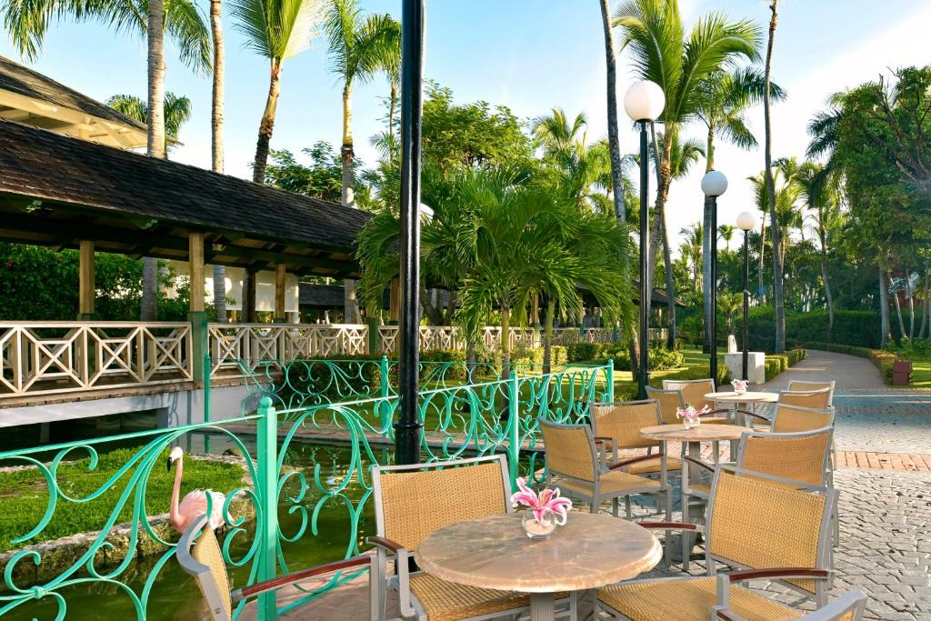 Trokšņains atpūtas laukums ar galdiem un krēsliem, ieskauts tropisko palmu un zaļumu. Daba un arhitektūra harmoniski apvienojas, radot mierīgu atmosfēru.