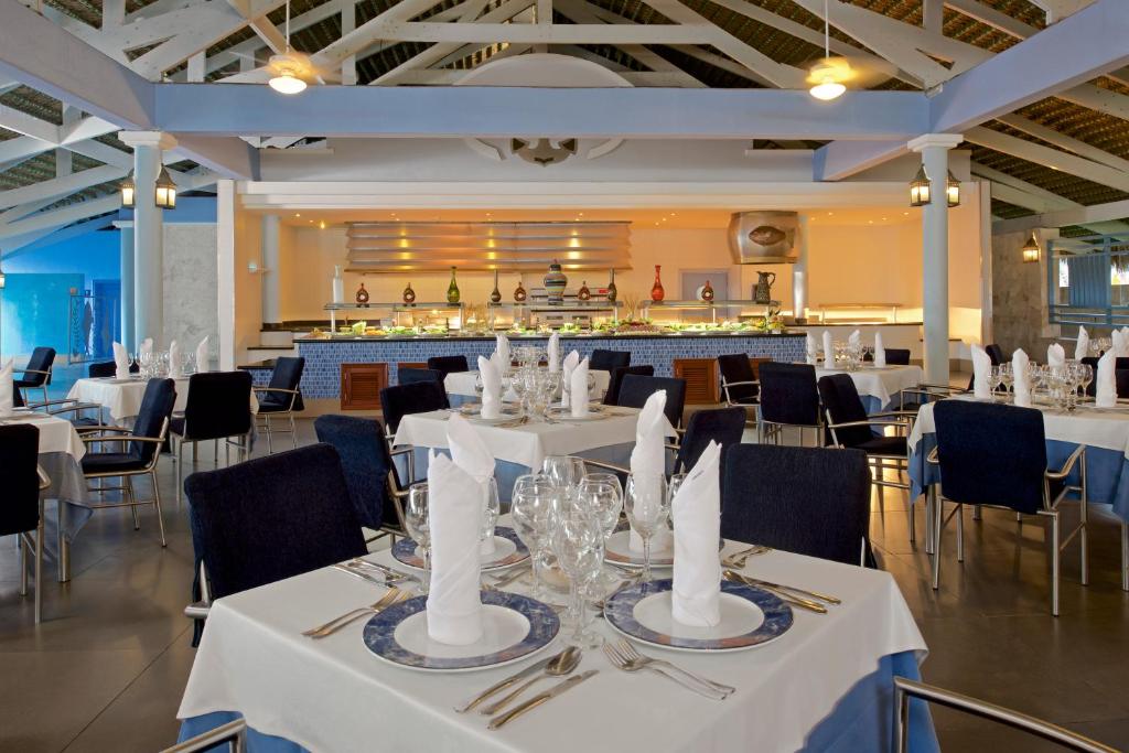 Moderna restorāna iekārtojums ar gaišām krāsām un elegantām galdniecēm, kas klāt ar baltiem galdautiem un grezniem galda piederumiem. Fons veido atvērta bāra zona ar dekoratīvām pudelēm un patīkamu apgaismojumu.