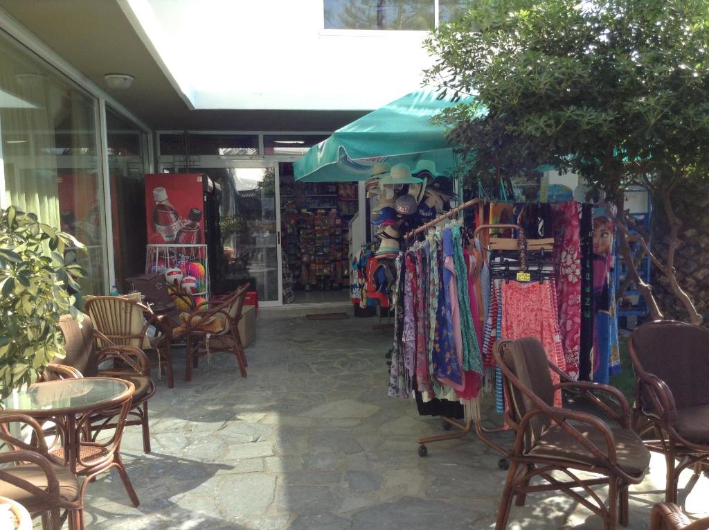 Veikala ieejas skats ar krāsainiem apģērbiem un aksesuāriem, kas izkārtoti uz pakaramajiem, un sēdvietām atpūtai. Apkārt ir augu zaļumi un sauļošanās telpa zem jumta.