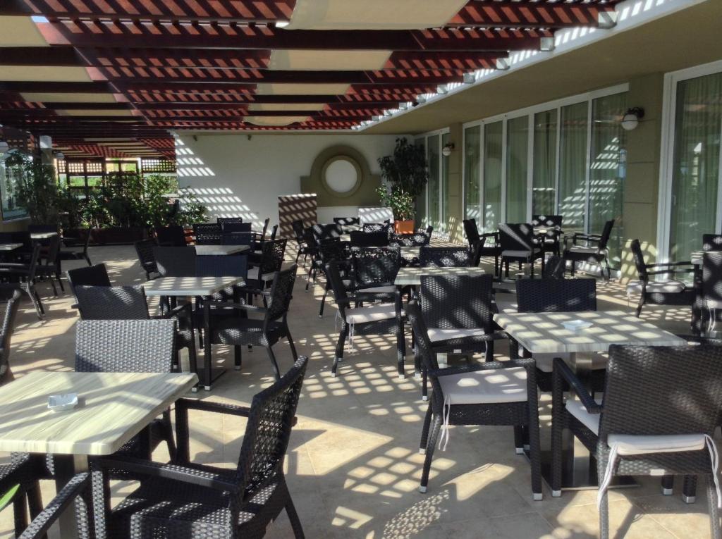 Ārējais restorāna terases skats ar melniem plastmasas galdiem un krēsliem, kas izvietoti zem jumta ar līstēm. Terase ir apzaļumota ar augiem un augu podiem.