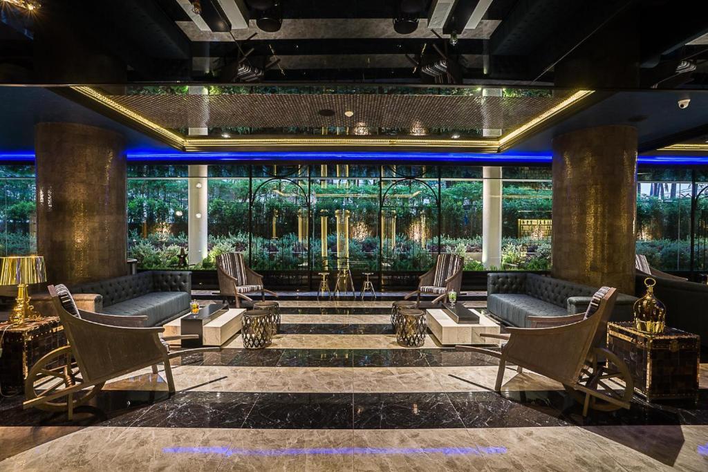 Mūsdienīga viesnīcas uzgaidāmā telpa ar elegantiem dīvāniem, stikla sienām un zaļo ainavu fonā. Interjerā ir izmantoti melni un zeltaini akcenti, radot izsmalcinātu atmosfēru.