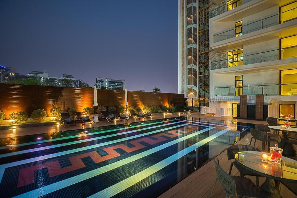 Luksusa viesnīcas baseins ar modernu dizainu un apgaismojumu naktī, ap kuru izvietoti sauļošanās krēsli. Fonā redzami mūsdienīgi pilsētas ēku silueti.