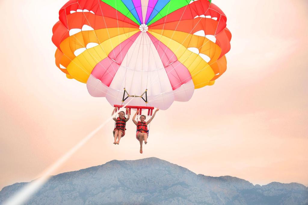 Divi cilvēki lido ar krāsainu parasail uz fona ar kalniem un saulainu debesu ainavu.