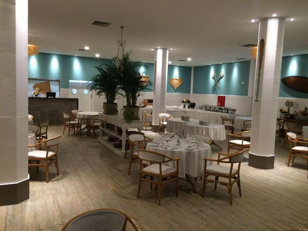 Mūsdienīgs restorāna interjers ar gaišām krāsām, elegantiem galdiem un koka krēsliem, kas papildināts ar dekoratīvām augu kompozīcijām un mākslinieciskām detaļām uz sienām.