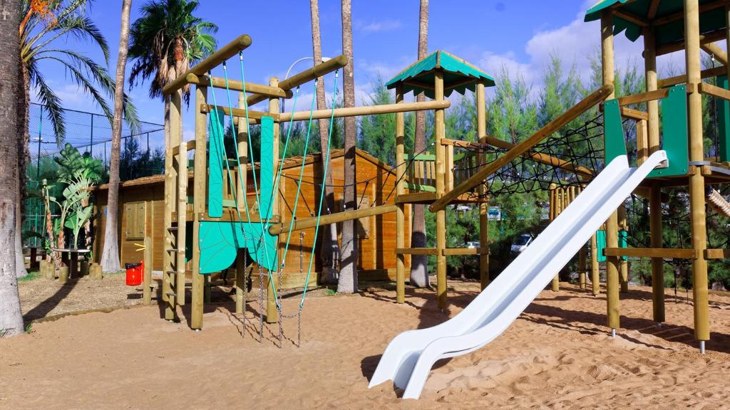 Bērnu spēļu laukums ar koka struktūrām, šūpolēm un slidkalniņu, apkārt palmām un zaļumiem. Ideāla vieta bērnu atpūtai un spēlēm.