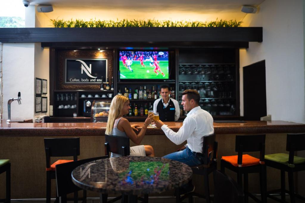 Elegants bārs ar modernu interjeru, kur pāris izbauda dzērienus, kamēr bārmenis apkalpo. Fons ar sporta pārraidi televizorā piešķir dzīvīgumu atmosfērai.