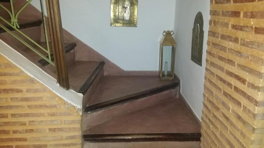 Koka kāpnes ar sarkanu apdari ved uz augšu, blakus ir dekoratīva laterna un mūra siena ar ķieģeļiem. Kāpņu telpa ir gaiša un mājīga, ar vienkāršu, bet stilīgu dizainu.