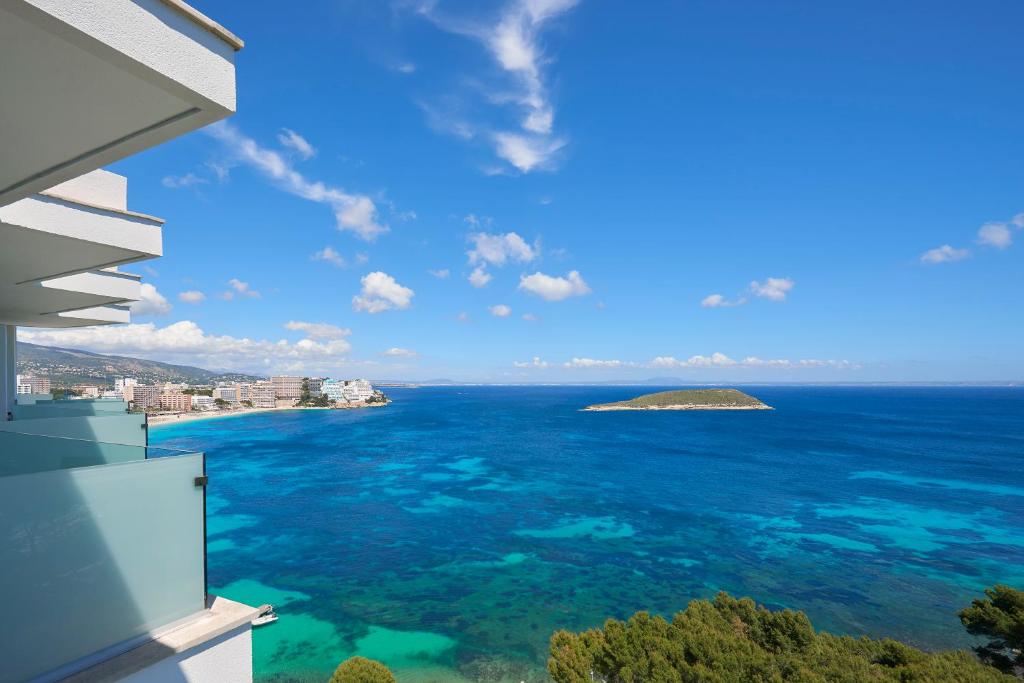 Skats no balkona, kur redzams skaidrs debesis un zilas jūras ūdeņi, kā arī neliela sala tālumā. Piekrastes pilsētas ainava un zaļumi piekrastē piešķir attēlam mierīgu un relaksējošu atmosfēru.