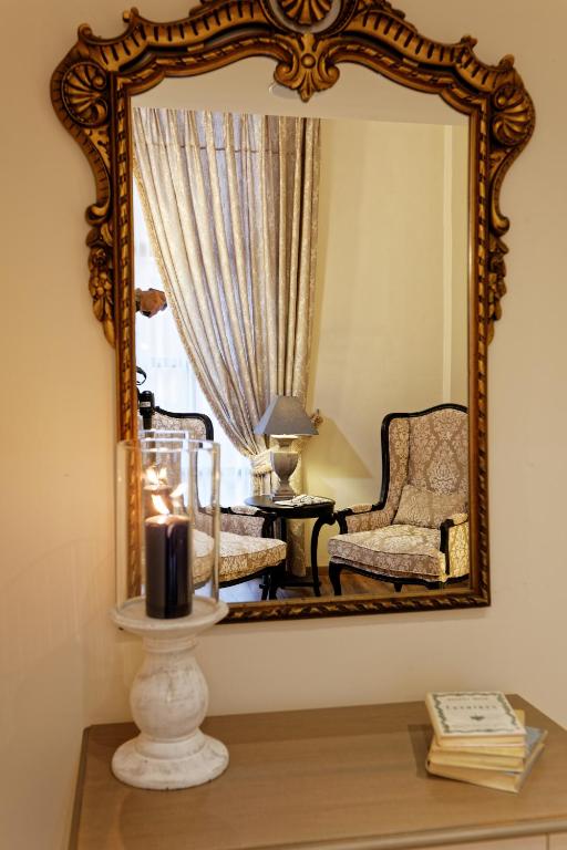 Elegants spogulis ar izsmalcinātu rāmi atspoguļo viesistabas interjeru, kurā redzami divi krēsli, galdiņš un aizkari. Attēlā ir arī svečturis ar sveci, kas piešķir siltumu un mājīgumu telpai.